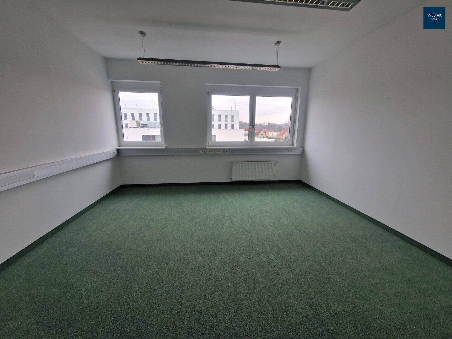 Leeres Büro mit Teppichboden, zwei Fenstern und Blick auf die umliegenden Gebäude.