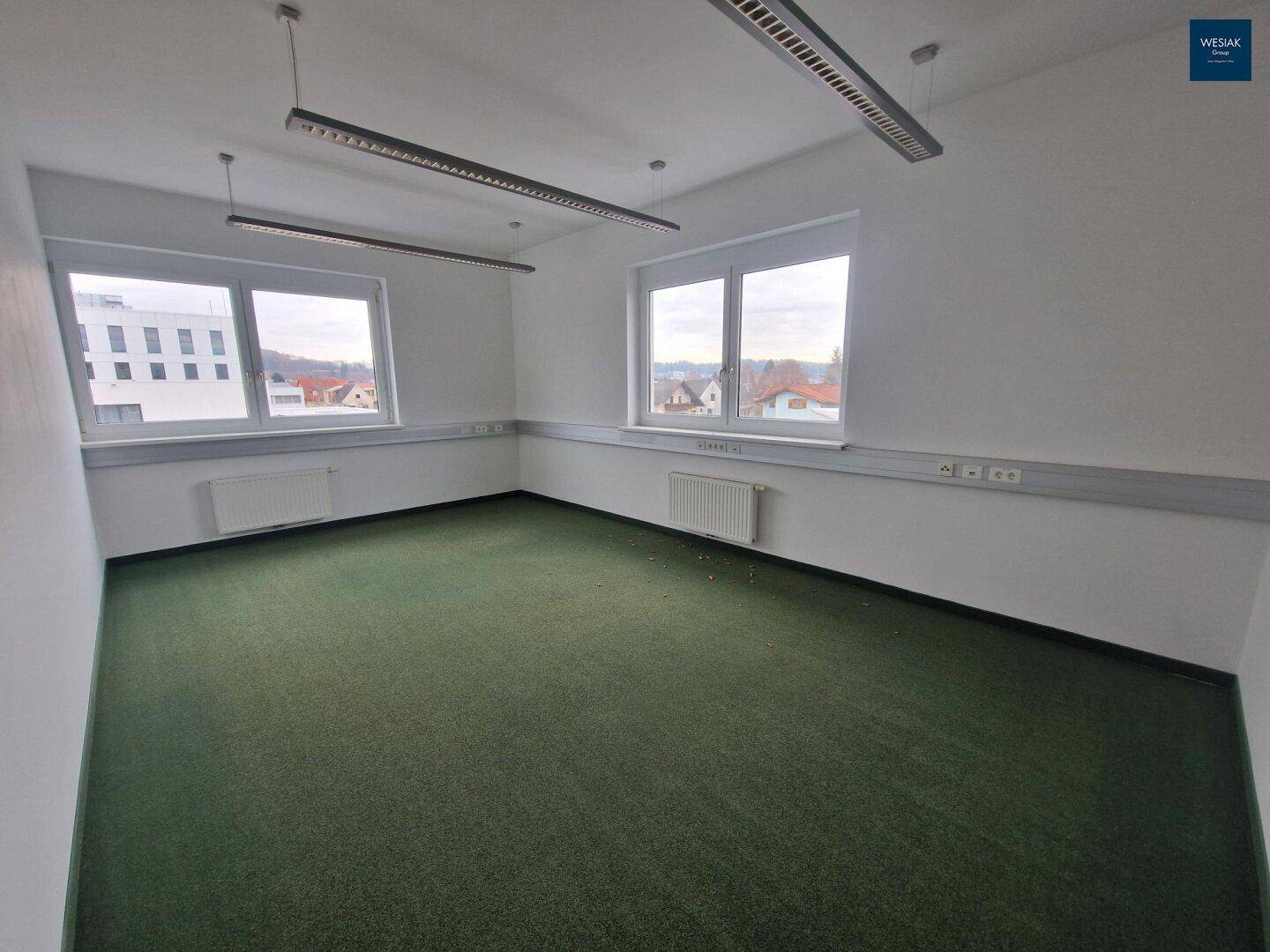 Leeres Büro mit Teppichboden, mehreren Fenstern und Blick auf die Stadt und Landschaft.