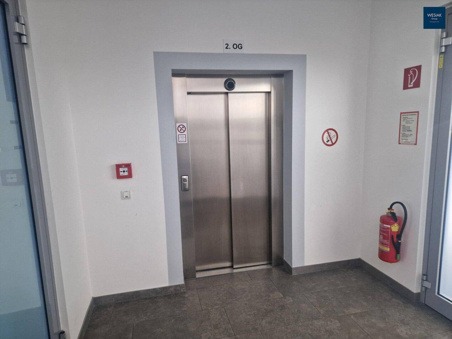 Aufzugsbereich mit Fliesenboden, Edelstahllift und Hinweisschildern im zweiten Obergeschoss.