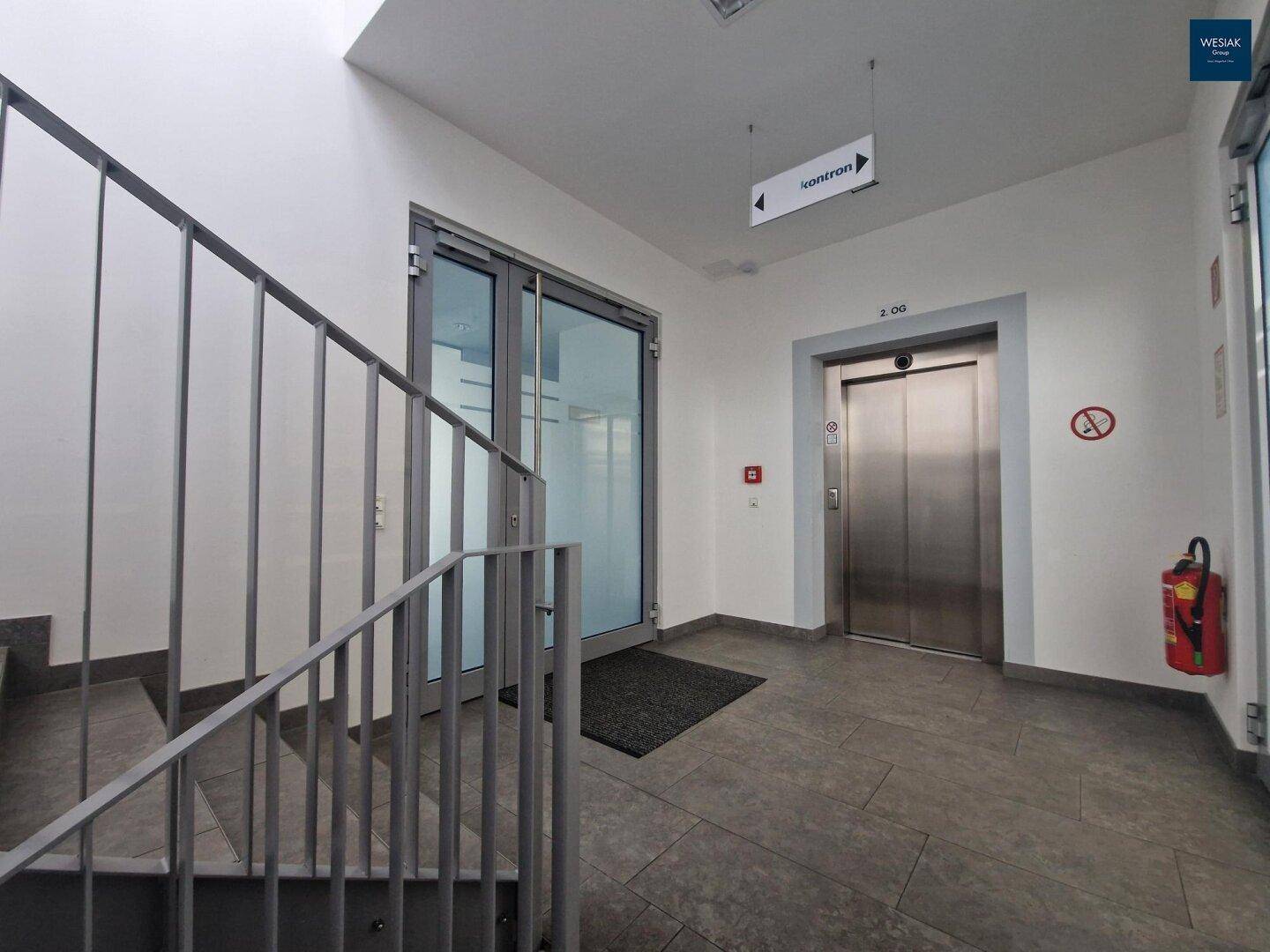 Eingangsbereich mit Treppe, Aufzug und Glasdoppeltür, die zu weiteren Räumen führt.
