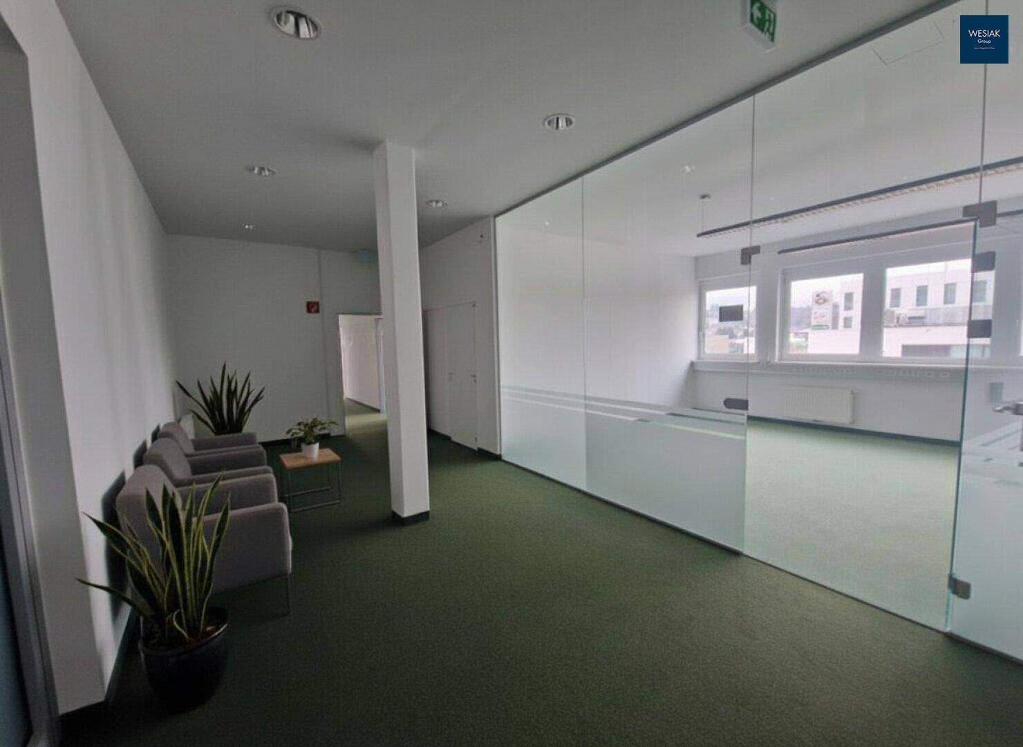 Wartebereich mit Teppichboden, bequemen Sesseln und einer Glaswand zum angrenzenden Büro.