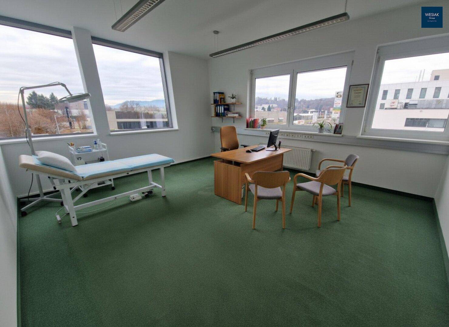Behandlungszimmer mit Teppichboden, Untersuchungsliege, Schreibtisch und großen Fenstern mit Ausblick.