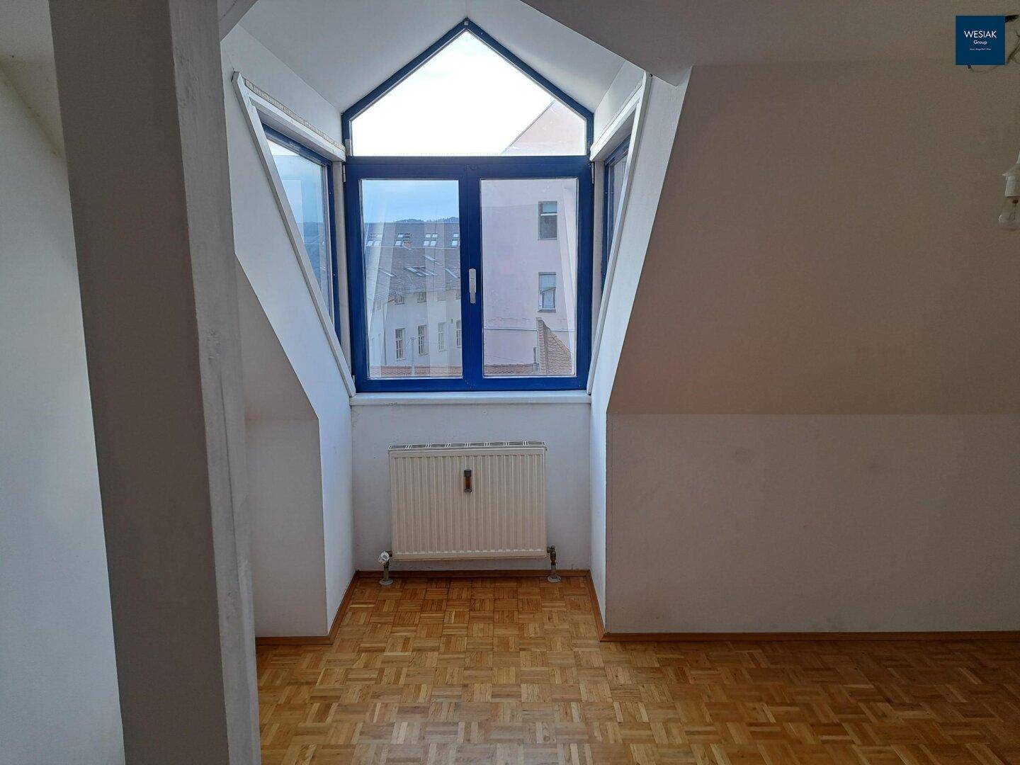 Heller Dachgeschossraum mit Parkettboden und charakteristischem Fenster, das viel Tageslicht hereinlässt.