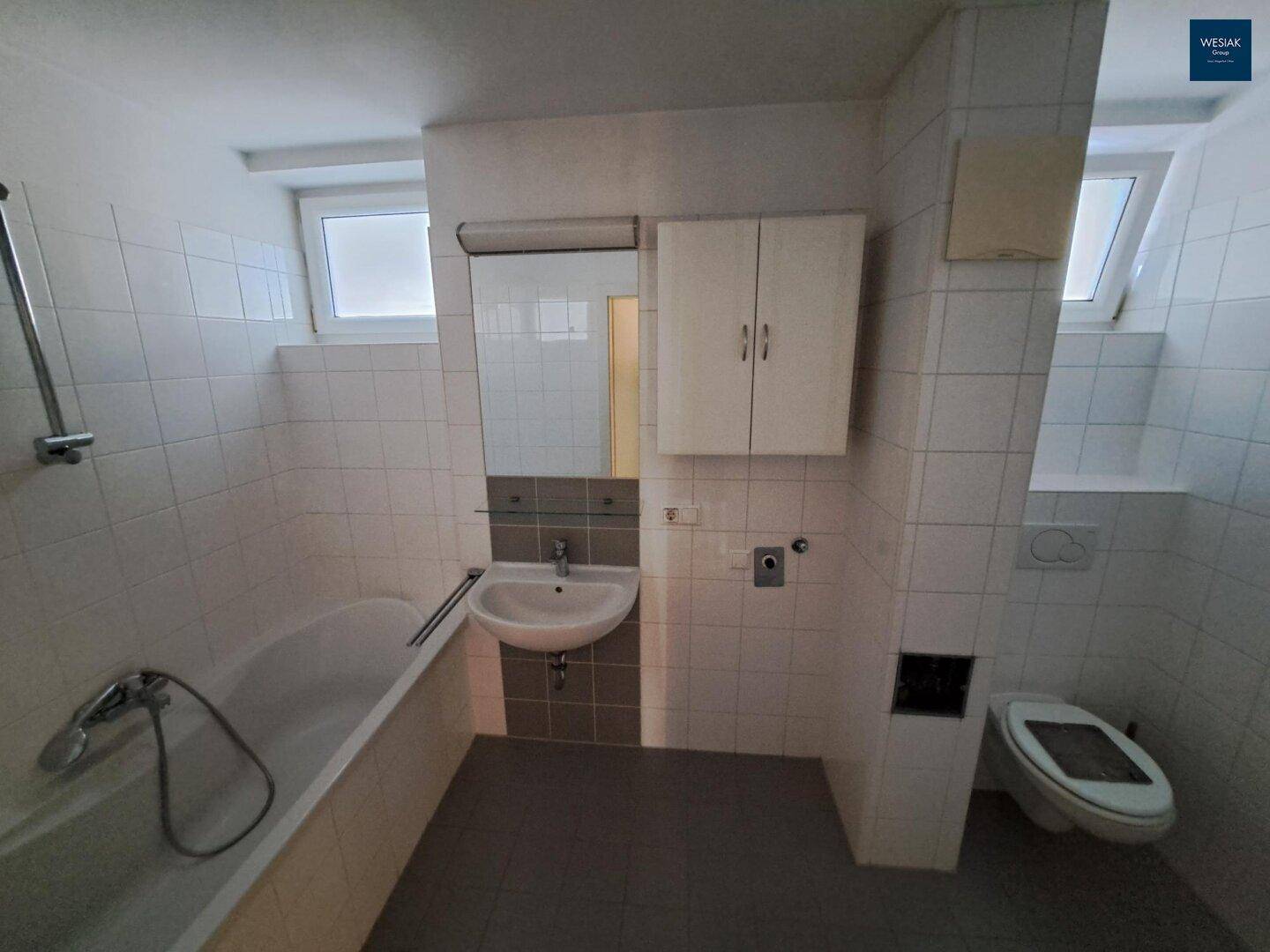 Gefliestes Badezimmer mit Badewanne, Waschbecken, Toilette und zwei Fenstern für Tageslicht.