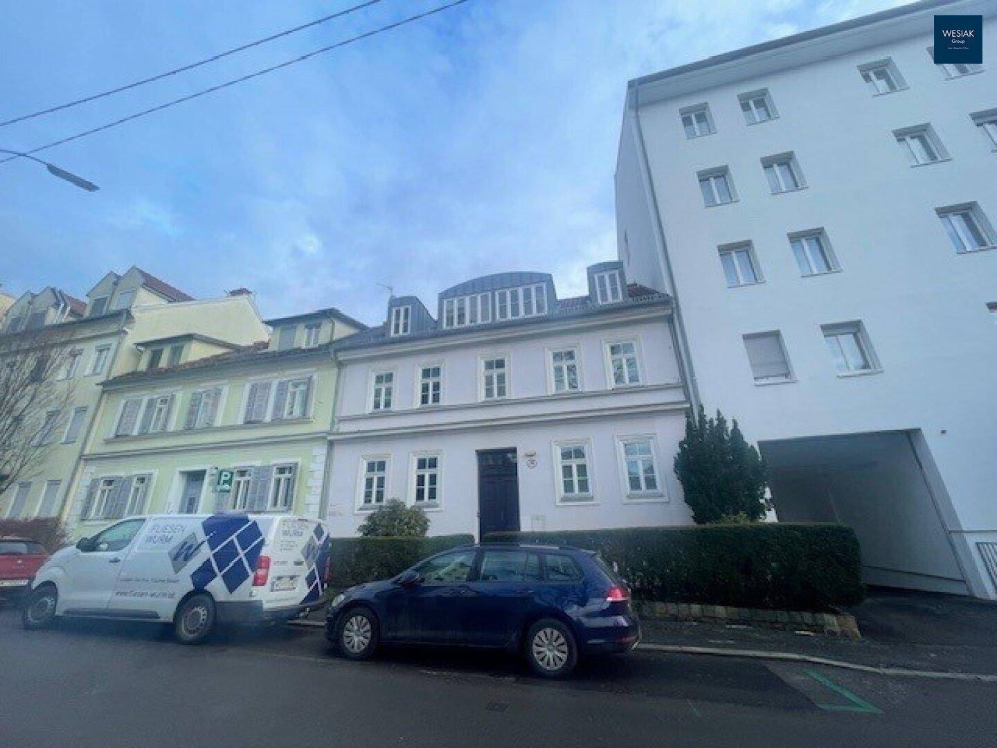 Gesamtansicht des Wohnhauses von der Straße mit angrenzenden Gebäuden und parkenden Autos.
