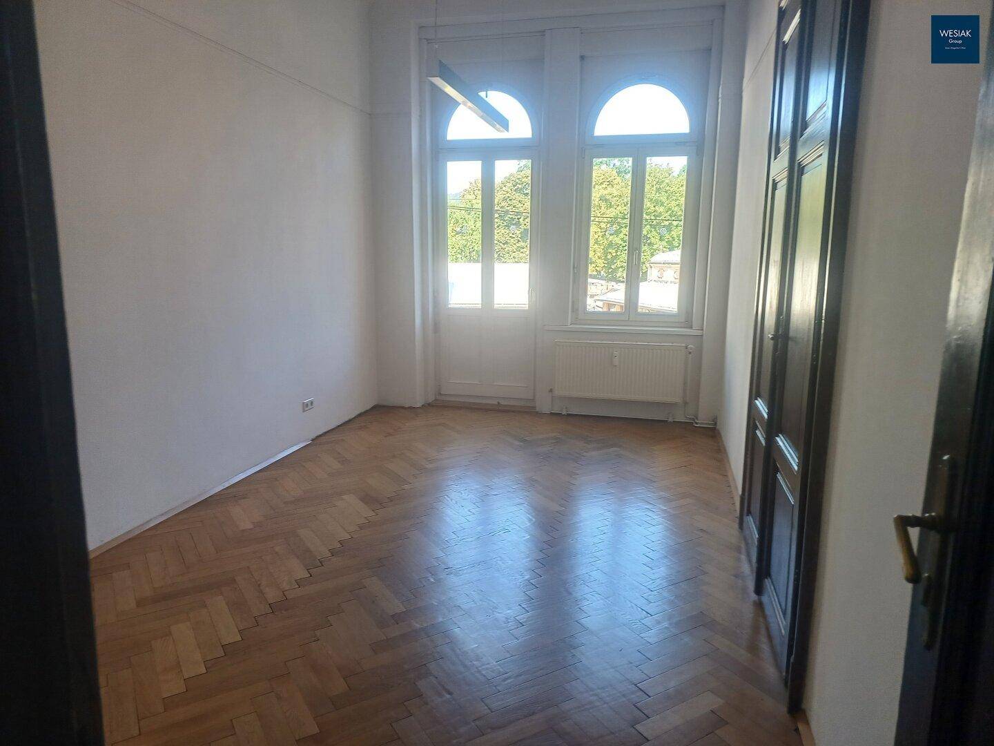 Helles Zimmer mit Fischgrätparkett und hohen Fenstern, die viel Tageslicht hereinlassen.