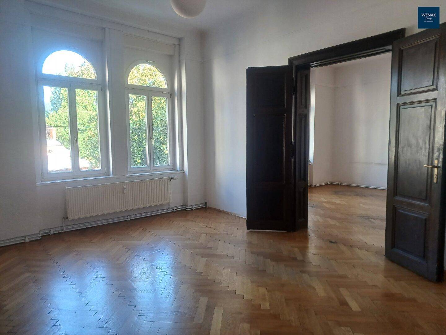 Geräumiges Zimmer mit klassischem Fischgrätparkett und hohen Fenstern, verbunden mit einem weiteren Raum.