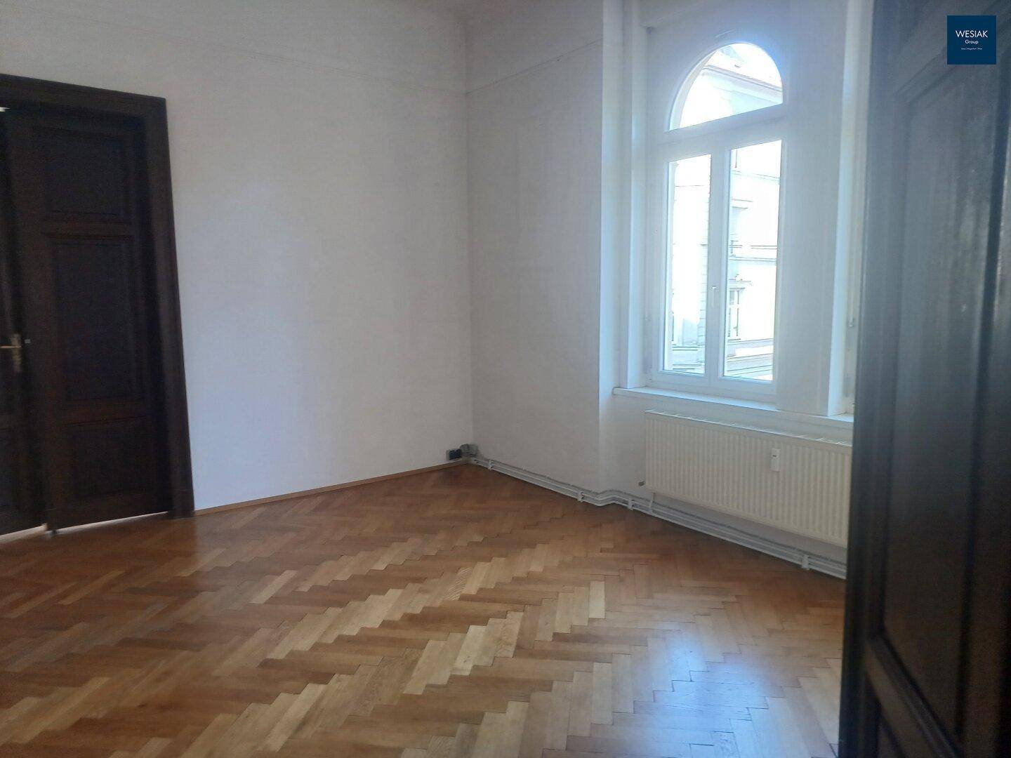 Lichtdurchflutetes Zimmer mit Fischgrätparkett und einem großen Bogenfenster, das den Raum erhellt.
