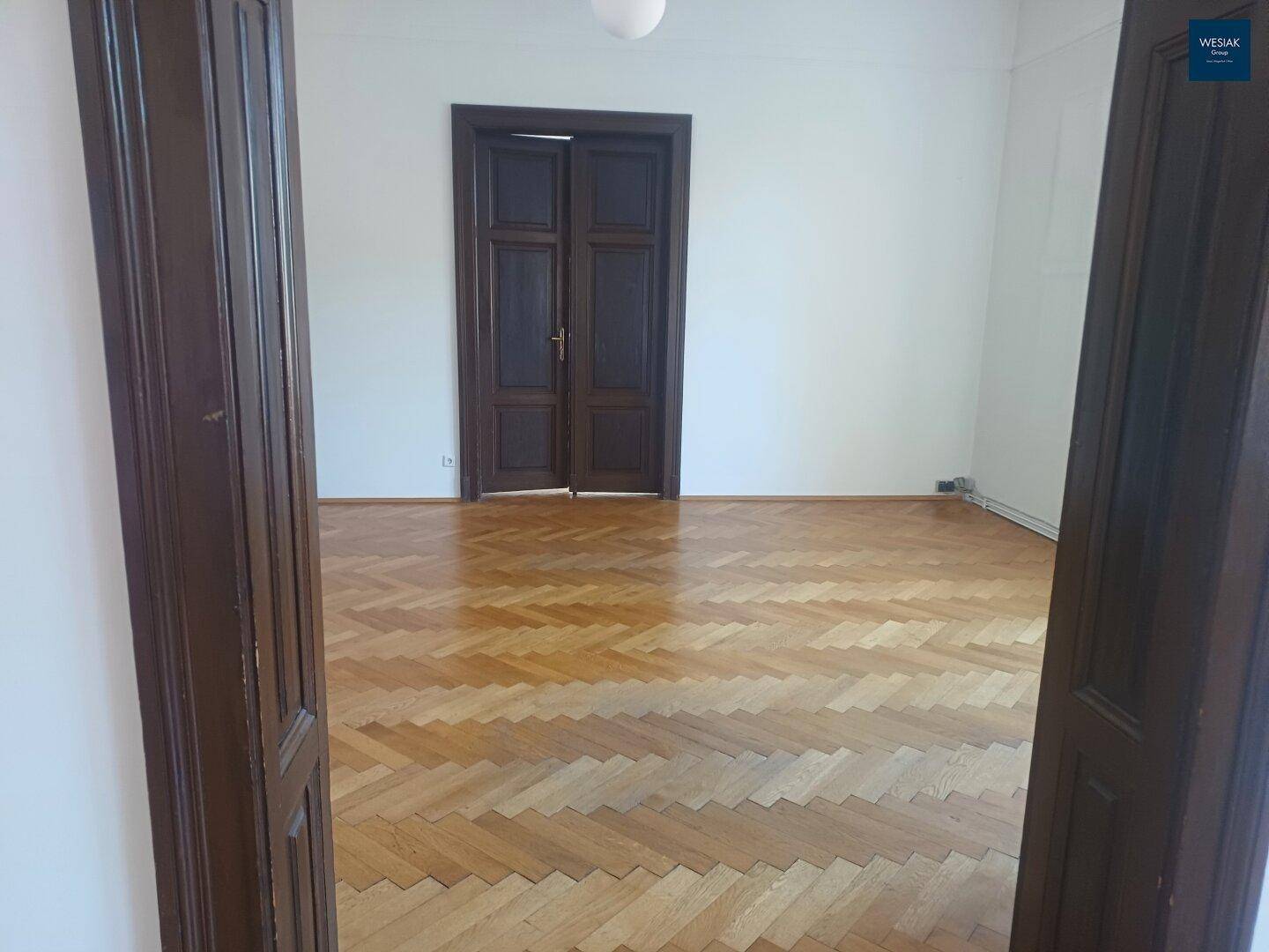 Leeres Zimmer mit Fischgrätparkett und hohen Doppeltüren, die den klassischen Stil des Altbaus betonen.