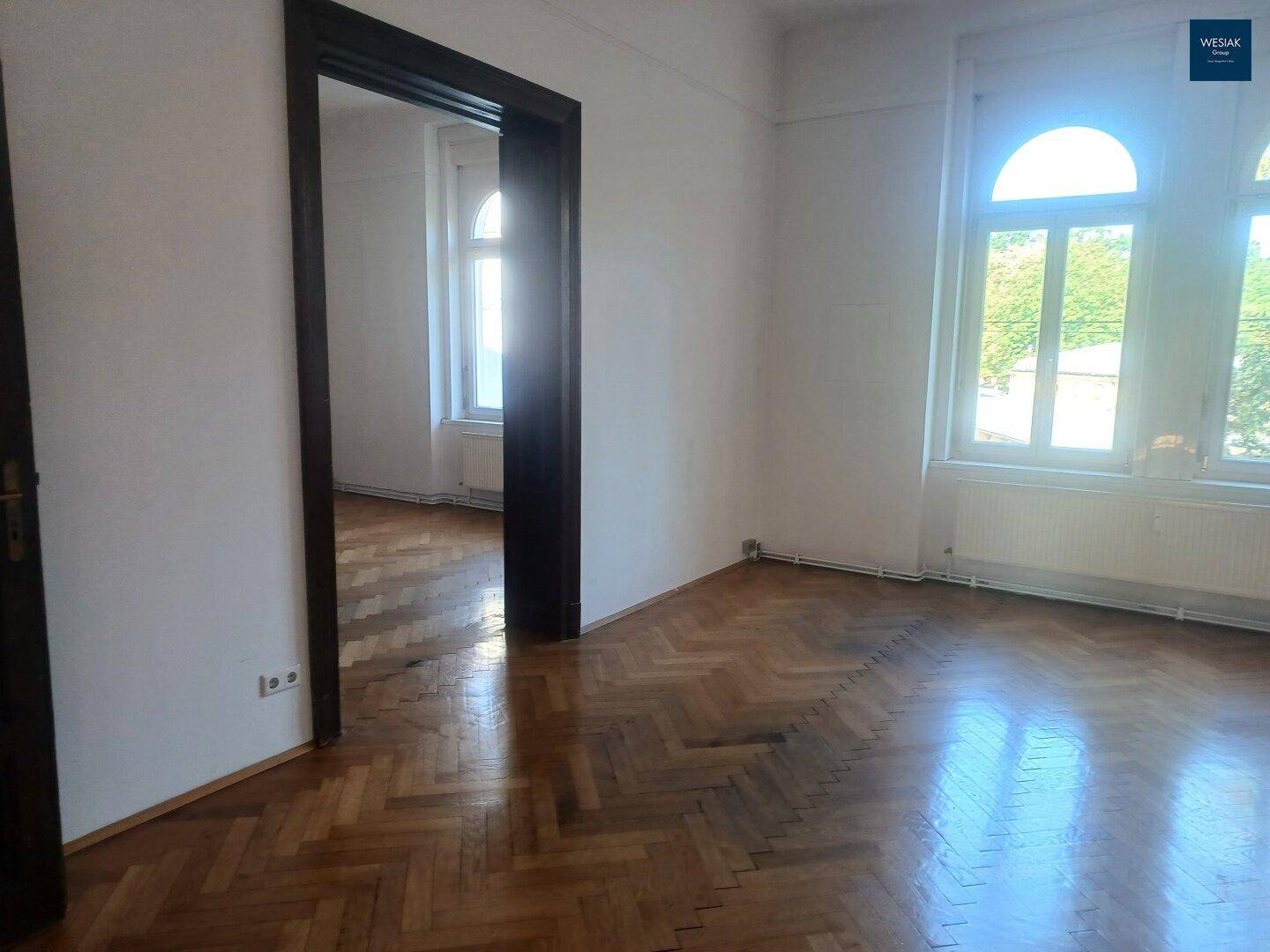 Helles Zimmer mit Fischgrätparkett und hohen Fenstern, die einen Blick ins Grüne ermöglichen.