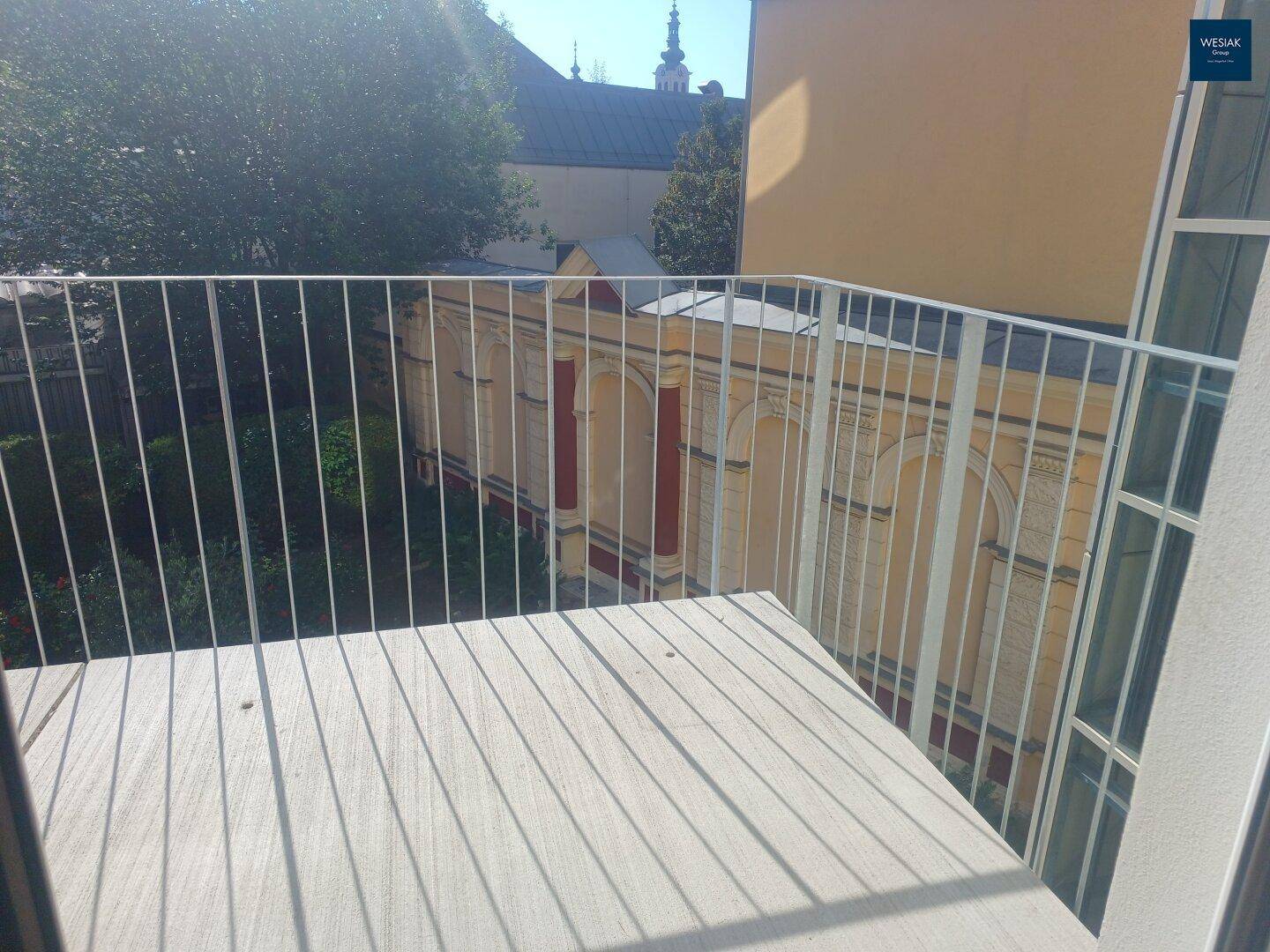 Sonniger Balkon mit Holzboden und Blick auf den grünen Innenhof und die umliegenden Gebäude.