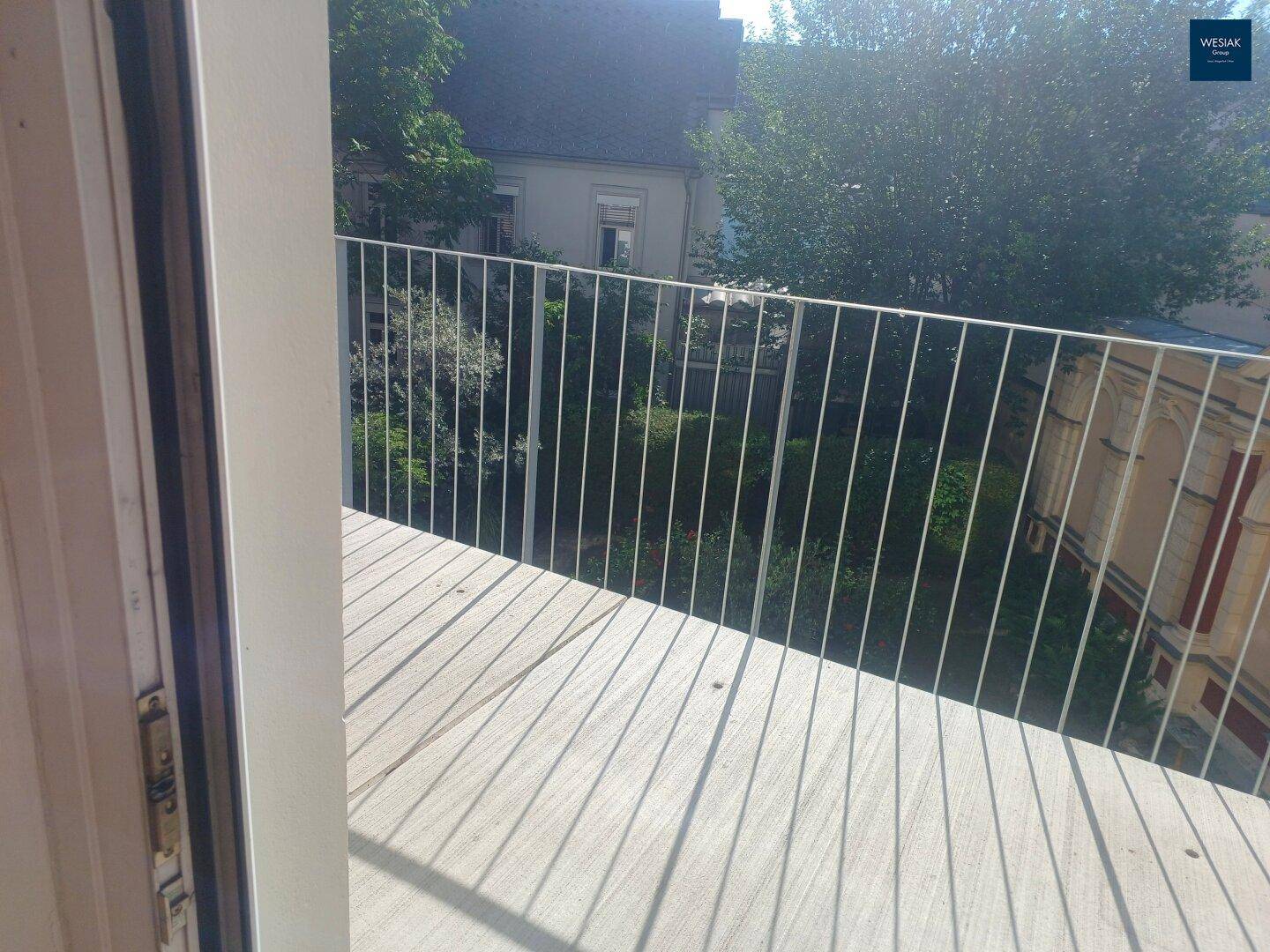 Heller Balkon mit Holzboden und weitem Blick über den grünen Innenhof und die Bäume.