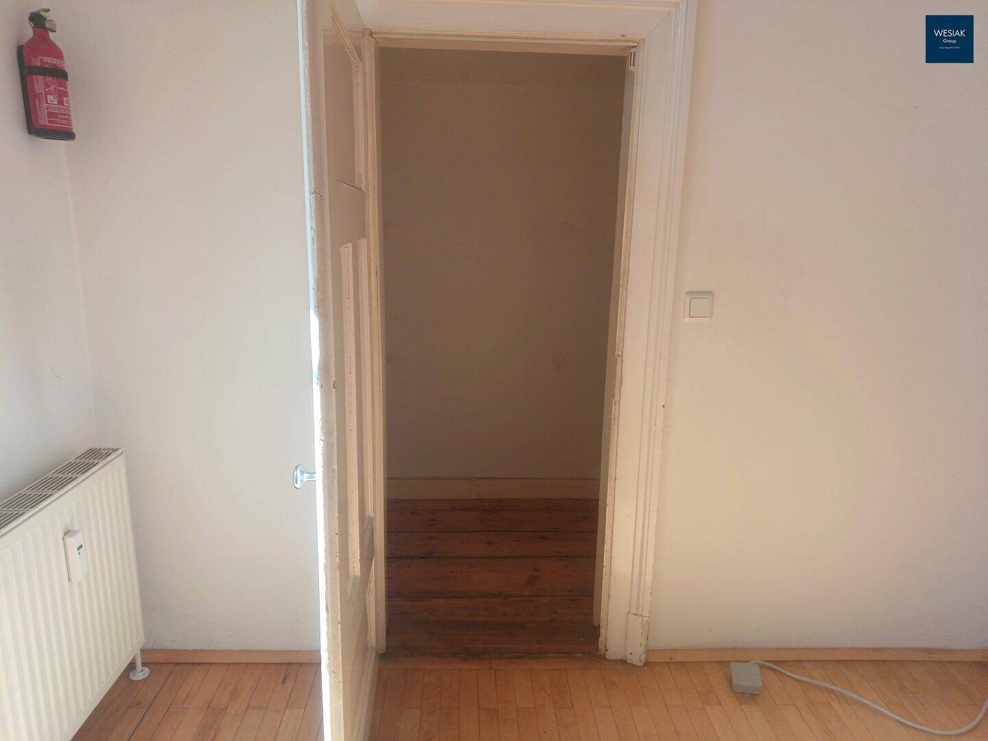 Blick durch eine offene Tür in einen leeren Raum mit Holzboden und Heizkörper.
