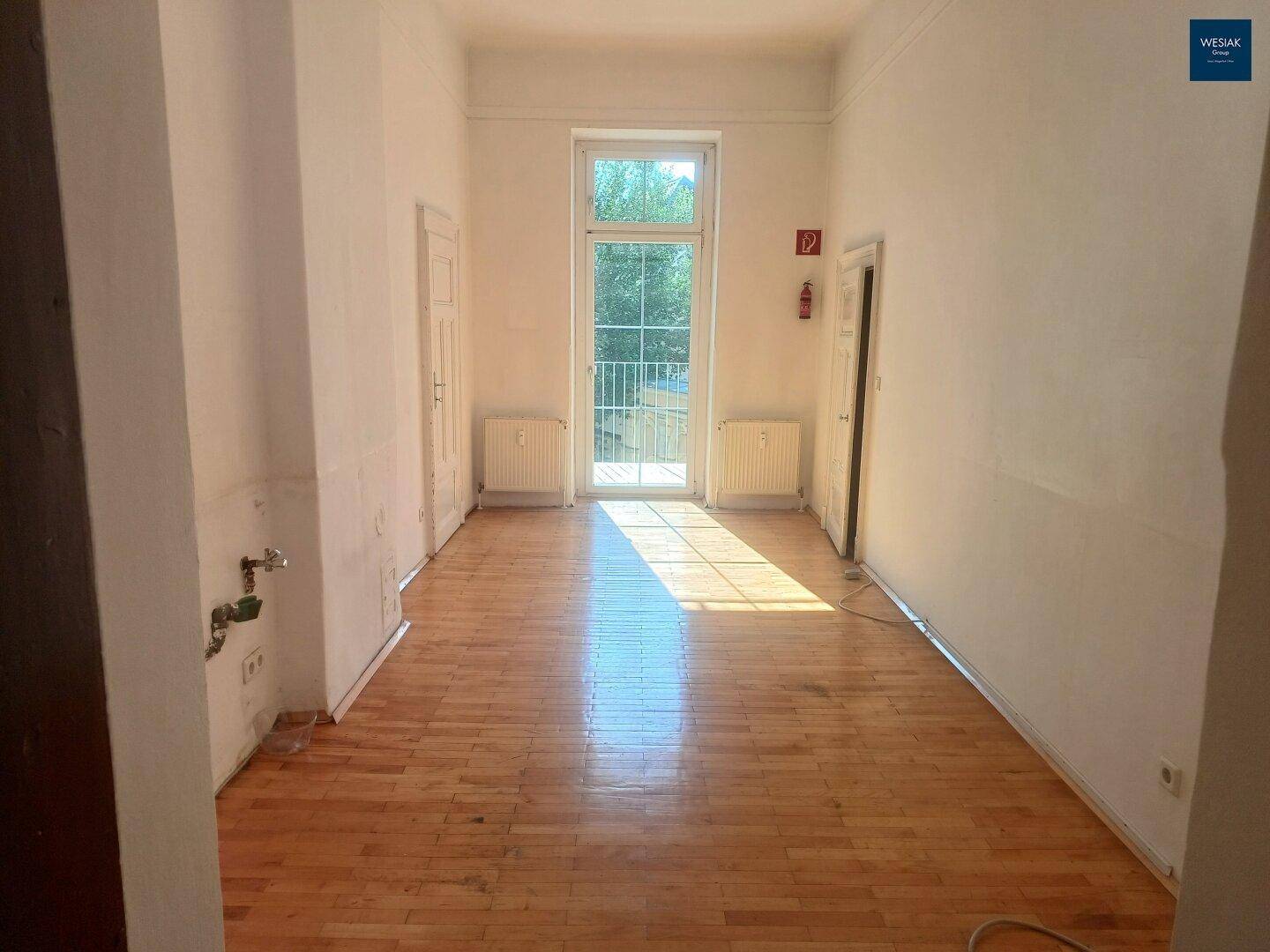 Geräumiger, leerer Wohnraum mit Fischgrätparkett, hohen Decken und französischem Balkon.