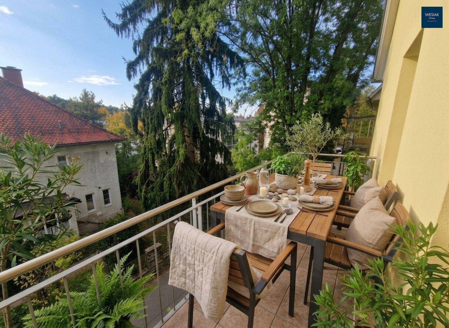 Gemütlicher Balkon mit Esstisch und Stühlen, umgeben von viel Grün und hohen Bäumen.