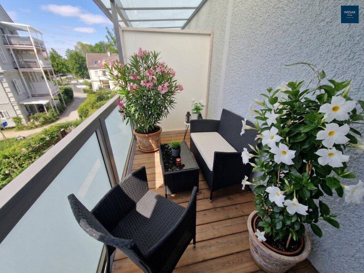 Gemütlicher Balkon mit Holzfußboden, Glasgeländer und blühenden Pflanzen, ideal zum Entspannen.
