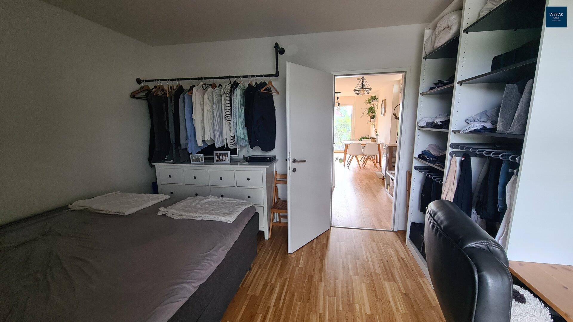 Schlafzimmer mit Bett, Kommode und offenem Kleiderschrank, mit Blick in den Wohnbereich.