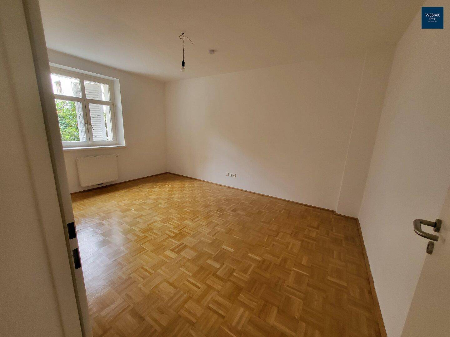 Helles Zimmer mit klassischem Parkettboden und einem Fenster mit Fensterläden.