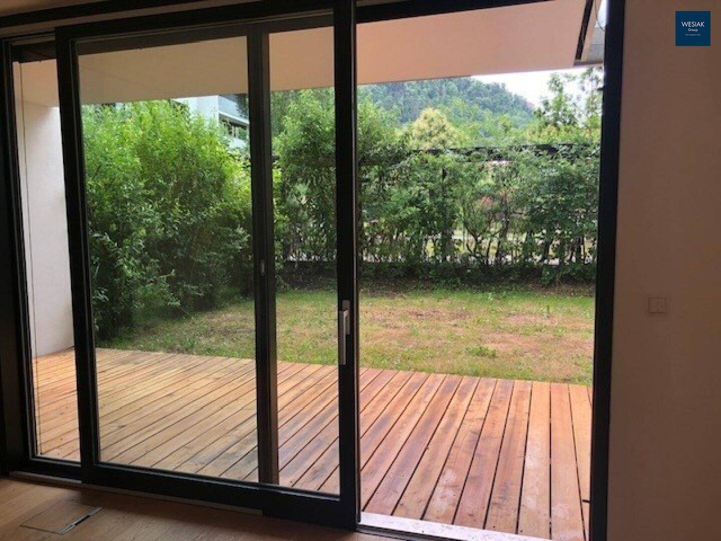 Große Glasschiebetüren bieten Zugang zu einer Holzterrasse und einem grünen Gartenbereich.