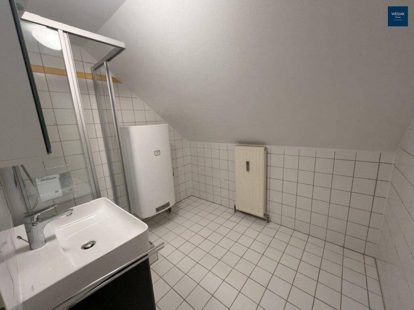Helles Badezimmer mit weißen Fliesen, Duschbereich und Dachschräge, bietet ausreichend Platz.