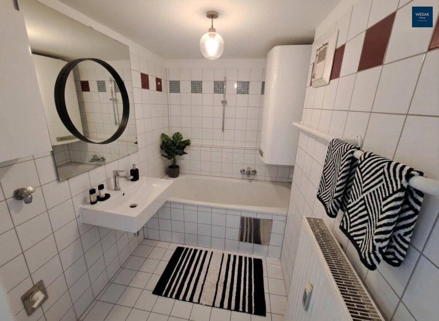 Gefliestes Badezimmer mit Badewanne, Waschbecken und Spiegel in Schwarz-Weiß-Optik.