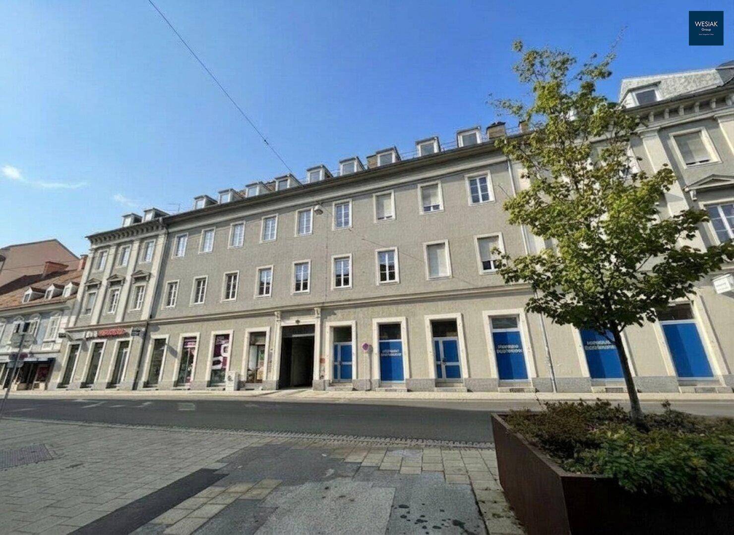 Historisches Gebäude mit grauer Fassade, Geschäften und einem Baum vor der Straße.