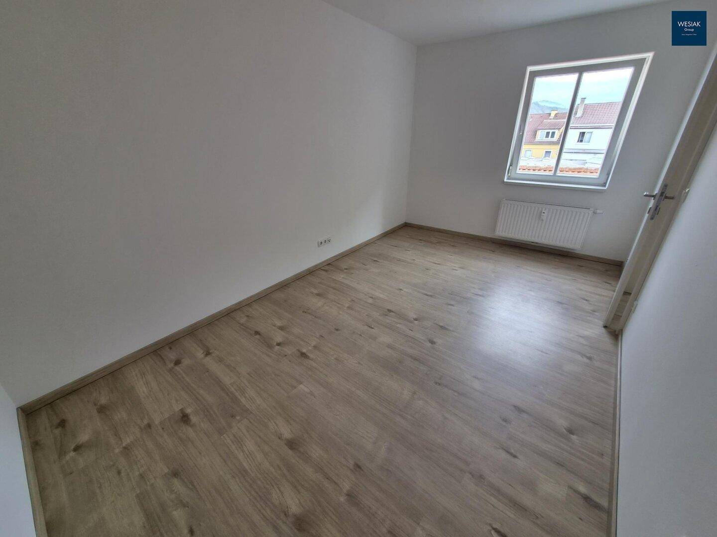 Helles, unmöbliertes Zimmer mit Laminatboden und einem Fenster mit Blick nach draußen.