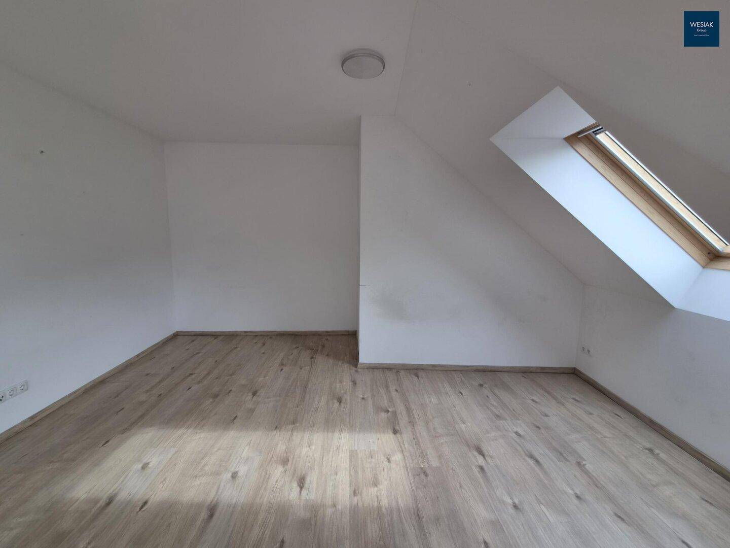 Leeres Zimmer im Dachgeschoss mit Laminatboden, weiß gestrichenen Wänden und Dachfenster.