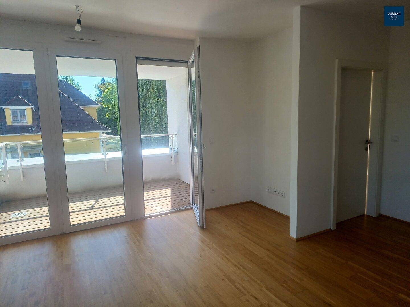 Heller Wohnraum mit Holzboden und Zugang zu einem Balkon mit Glasgeländer und Blick ins Grüne.