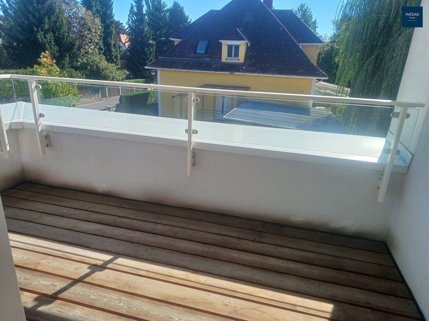 Balkon mit Holzbelag und Glasgeländer bietet einen angenehmen Blick auf die umliegende Bebauung und Grünflächen.