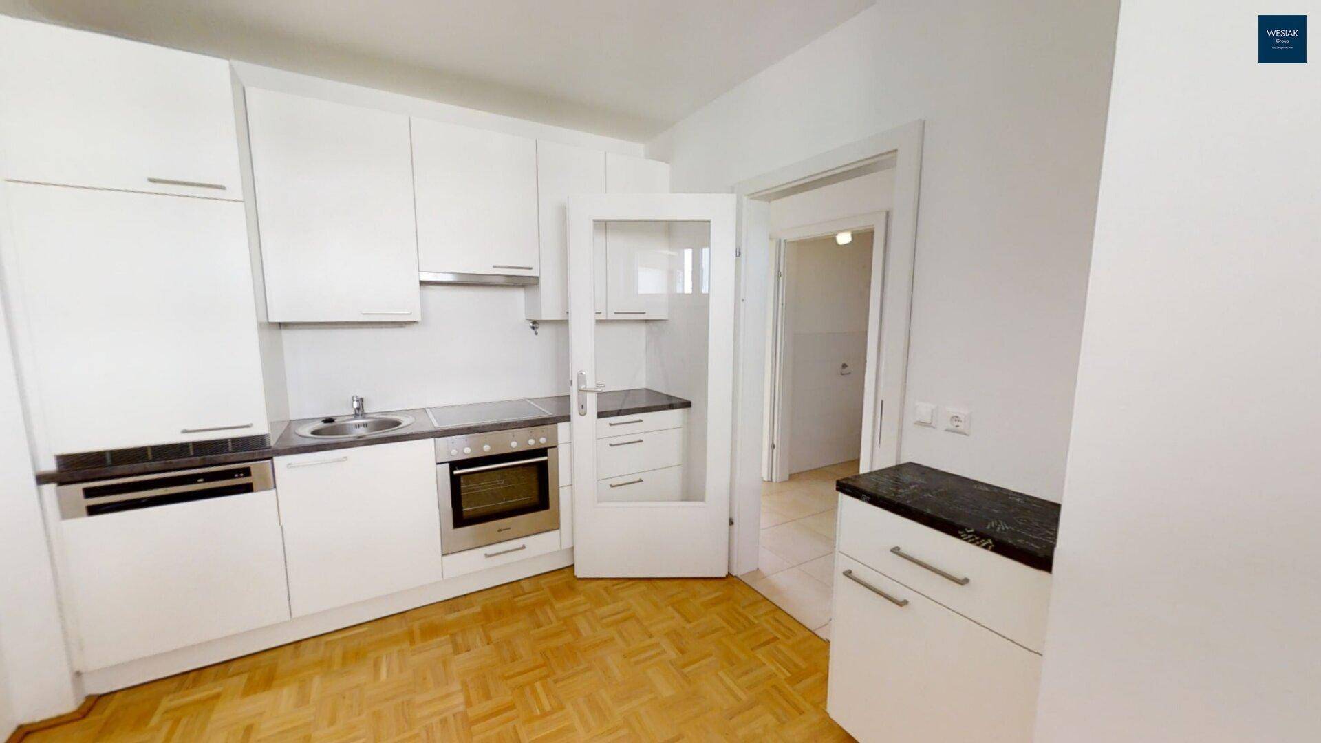 Moderne Einbauküche mit weißen Schränken, dunkler Arbeitsplatte und Parkettboden.