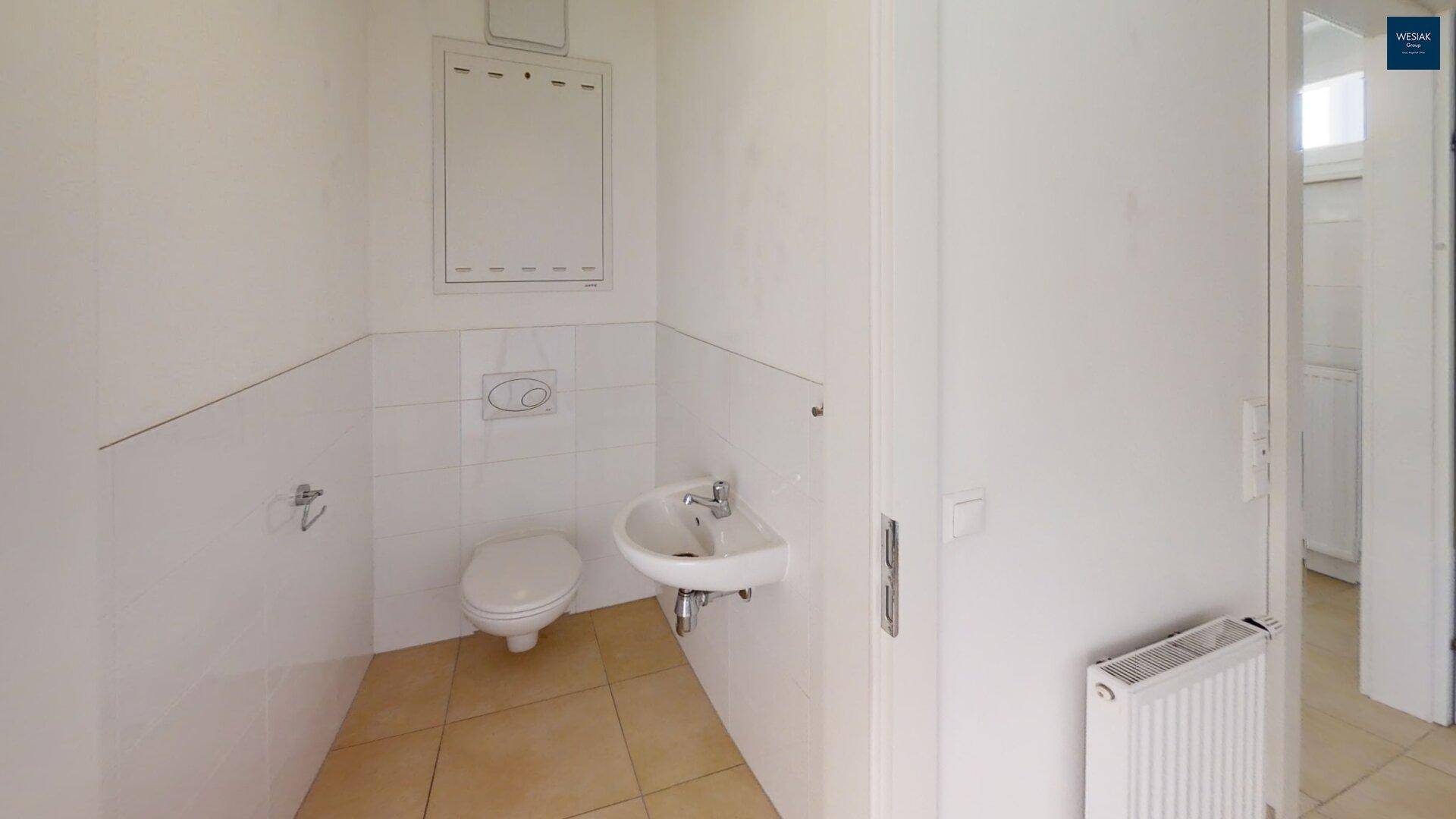 Separates WC mit Wand-WC, kleinem Waschbecken und hellen Fliesen.