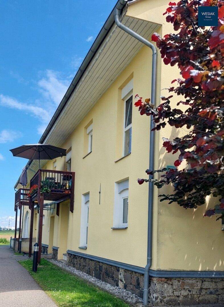 Seitenansicht des Gebäudes mit Balkon und umgebenden Bäumen unter blauem Himmel.