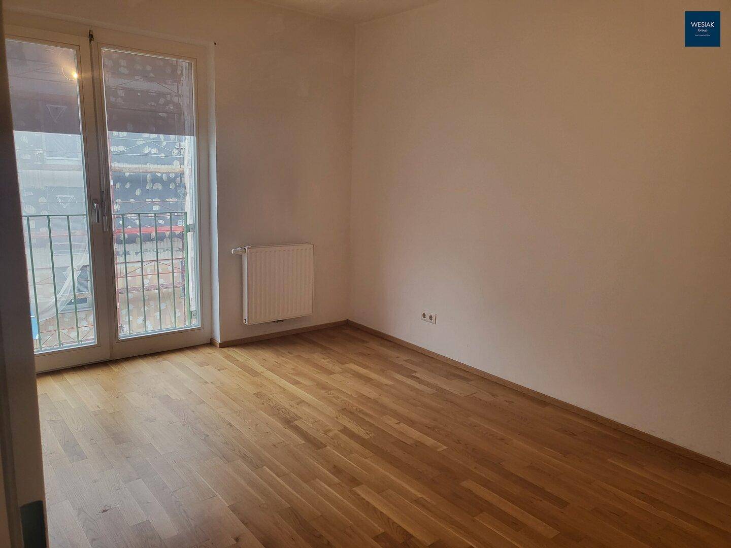 Lichtdurchfluteter Raum mit Parkettboden, Heizkörper und Zugang zu einem französischen Balkon.