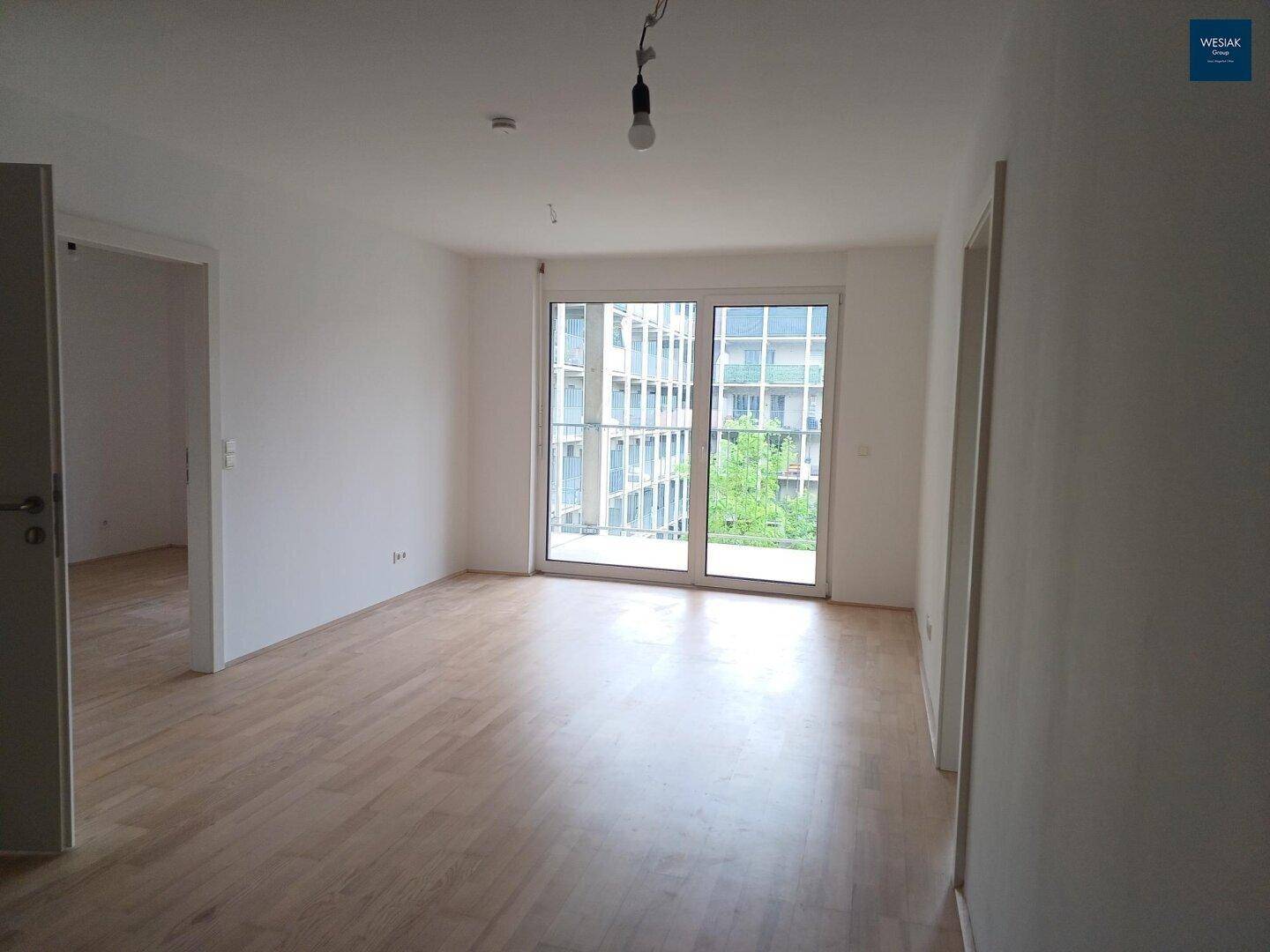 Helles, leeres Zimmer mit Parkettboden und großem Fenster, das Zugang zu einem Außenbereich bietet.