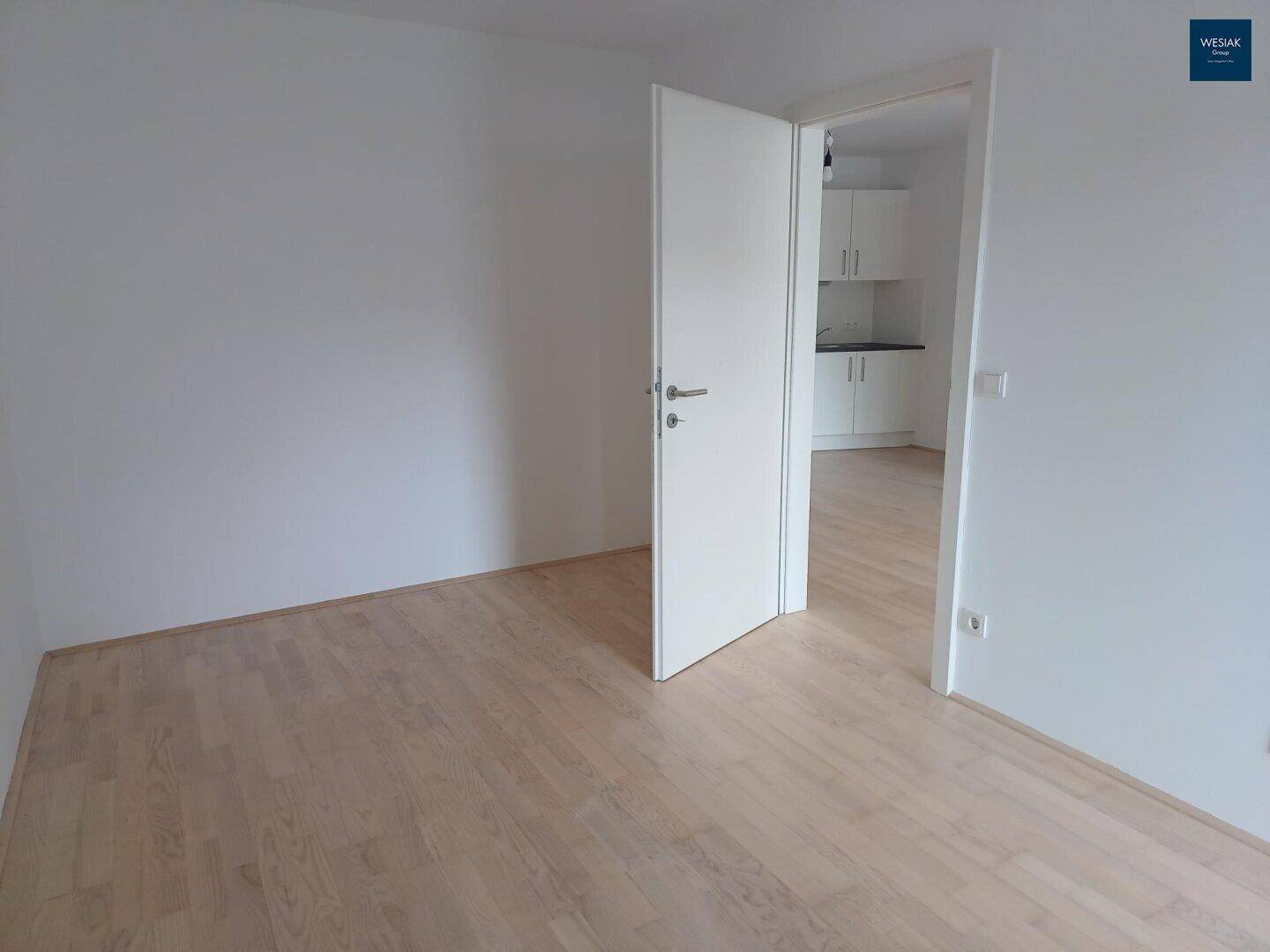 Leerer Raum mit Parkettboden und offener Tür, die den Blick in eine angrenzende Küche freigibt.