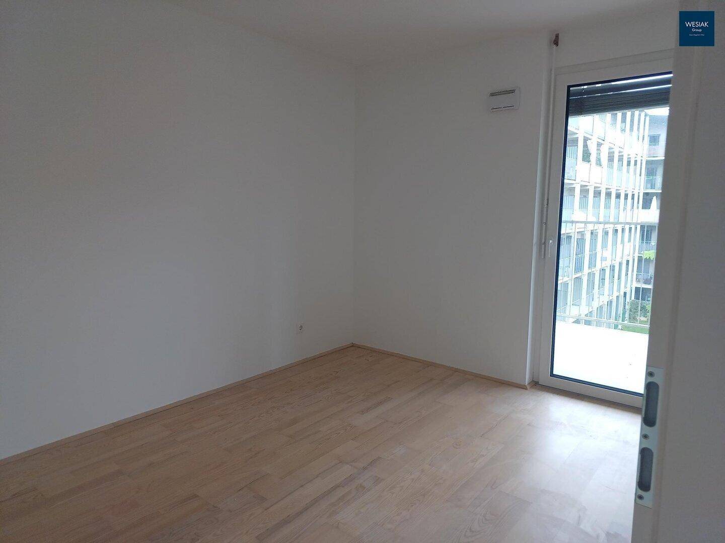 Helles, leeres Schlafzimmer mit Parkettboden und einem großen Fenster, das viel Tageslicht hereinlässt.
