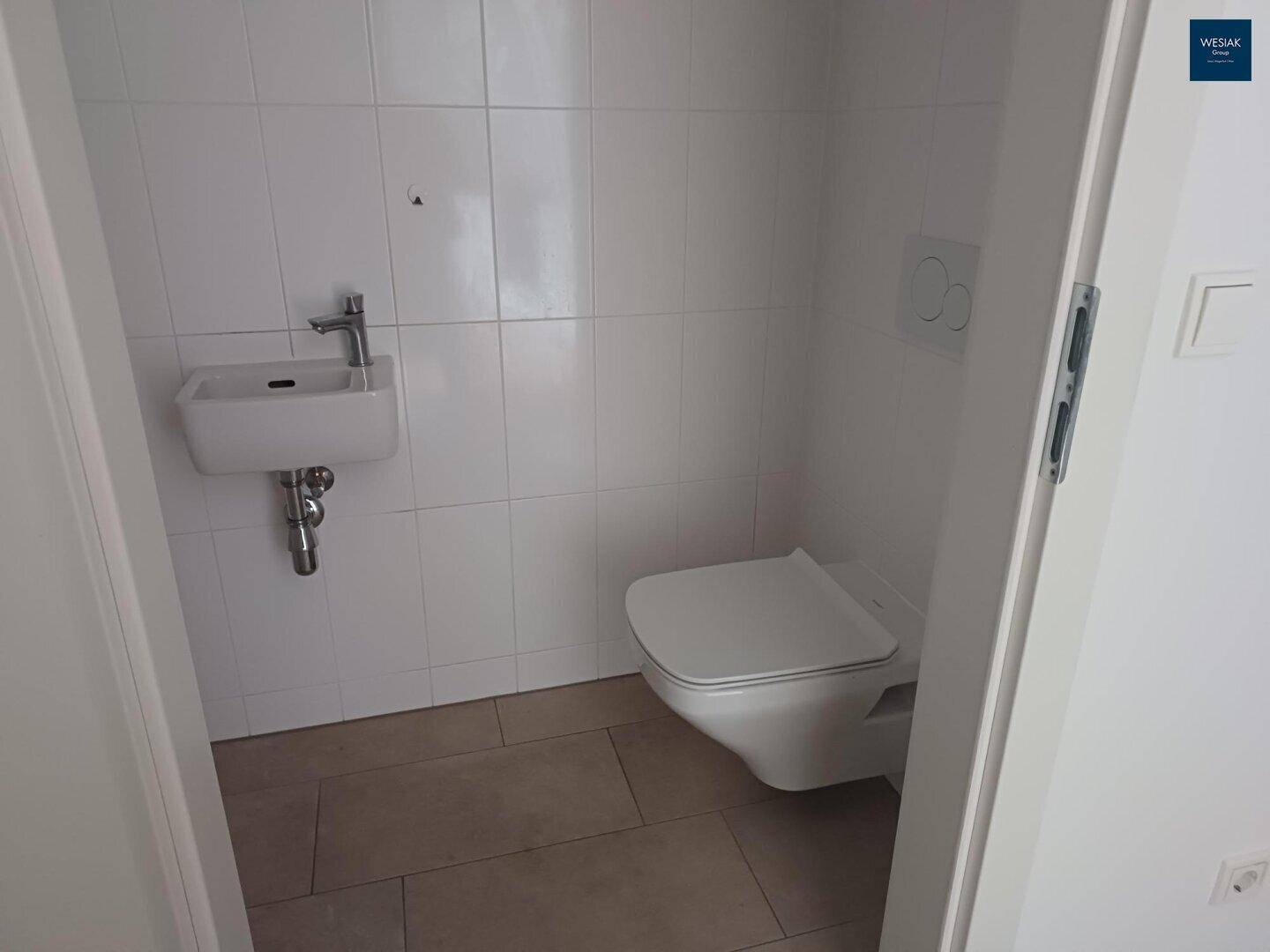 Separates Gäste-WC mit wandhängender Toilette, kleinem Waschbecken und hellen Fliesen an Wänden und Boden.