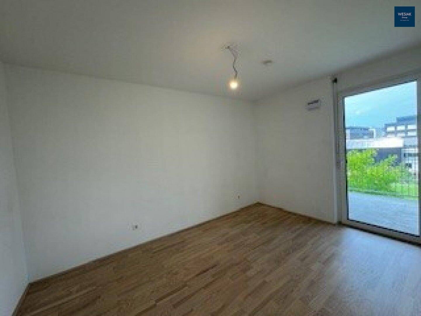 Geräumiges Schlafzimmer mit hellem Holzparkettboden und großem Fenster mit Zugang nach draußen.