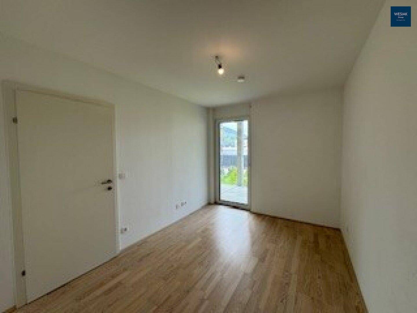 Helles Zimmer mit Holzfußboden und einem hohen Fenster, das viel Tageslicht hereinlässt.