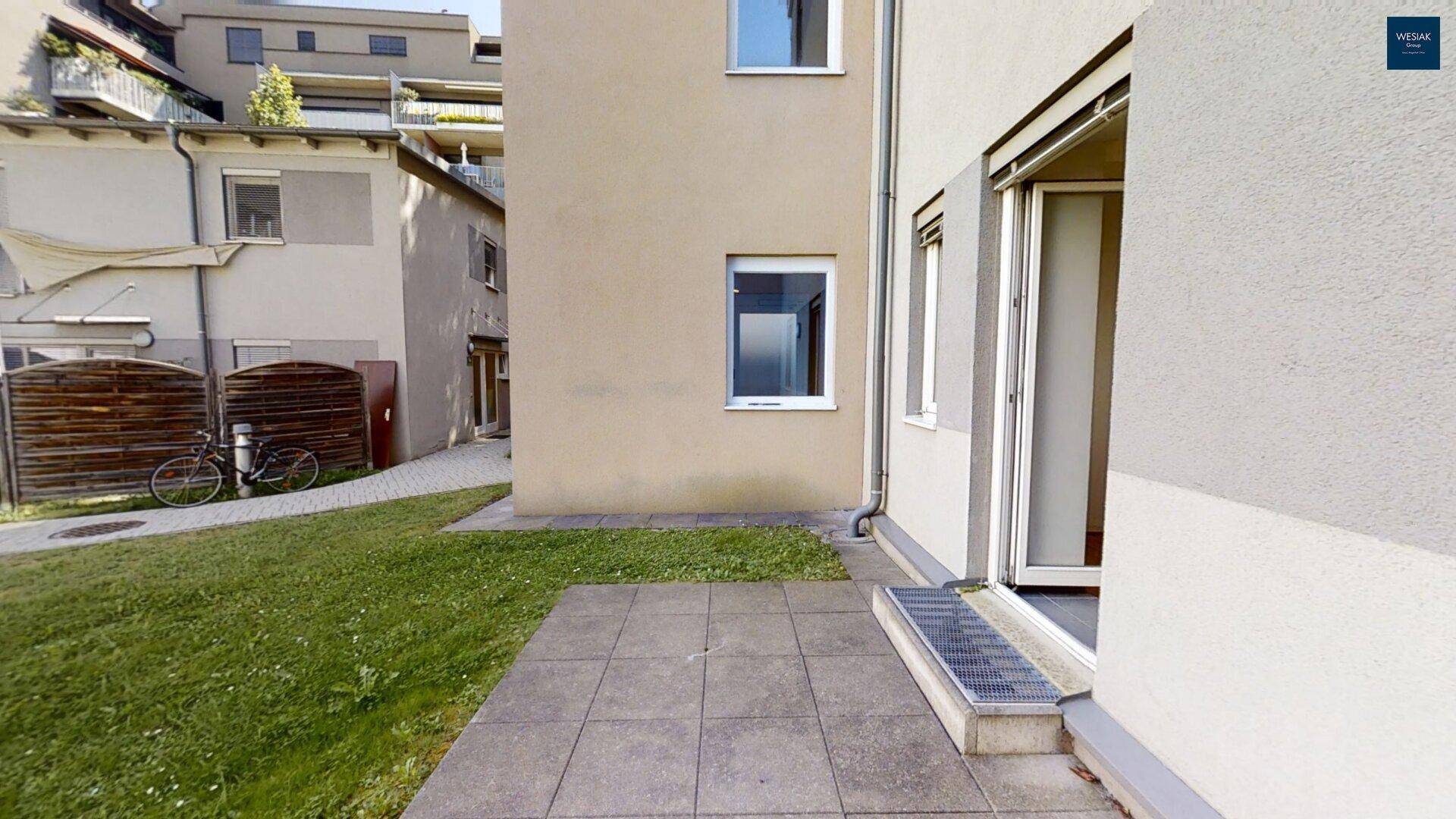 Gepflasterte Terrasse mit Rasenfläche und direktem Zugang zum Gebäude, umgeben von weiteren Wohnhäusern.