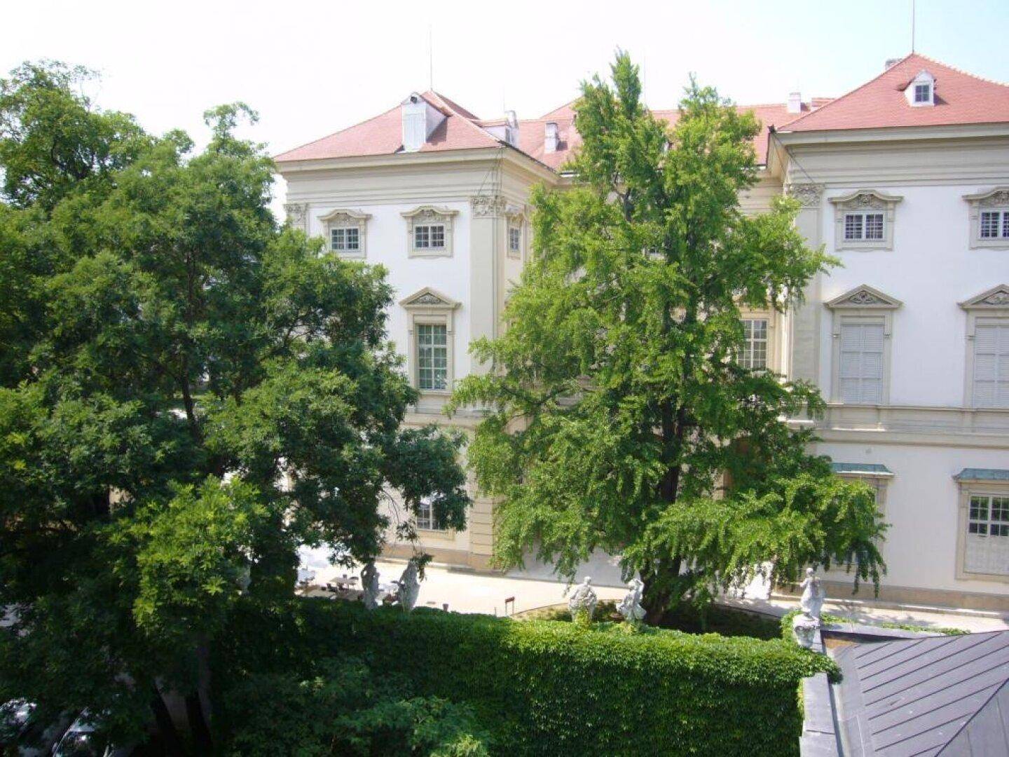 Blick auf ein historisches Gebäude mit gepflegten Bäumen und einer grünen Hecke im Vordergrund.