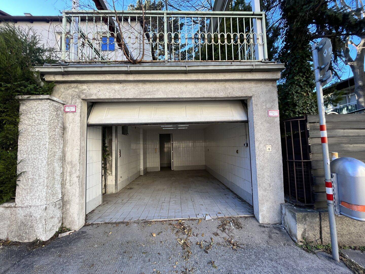 Außenansicht einer Garage mit geöffnetem Tor und darüberliegendem Balkon mit schmiedeeisernem Geländer.
