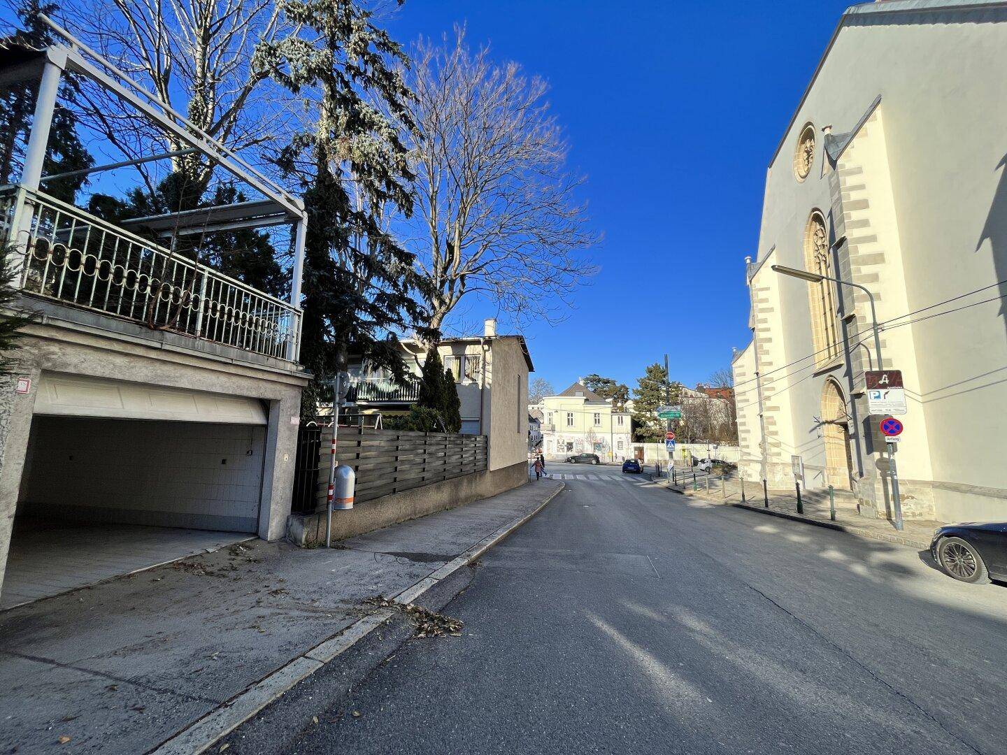 Sonniger Straßenblick mit dem Garagengebäude links, einer Kirche und weiteren Häusern unter blauem Himmel.