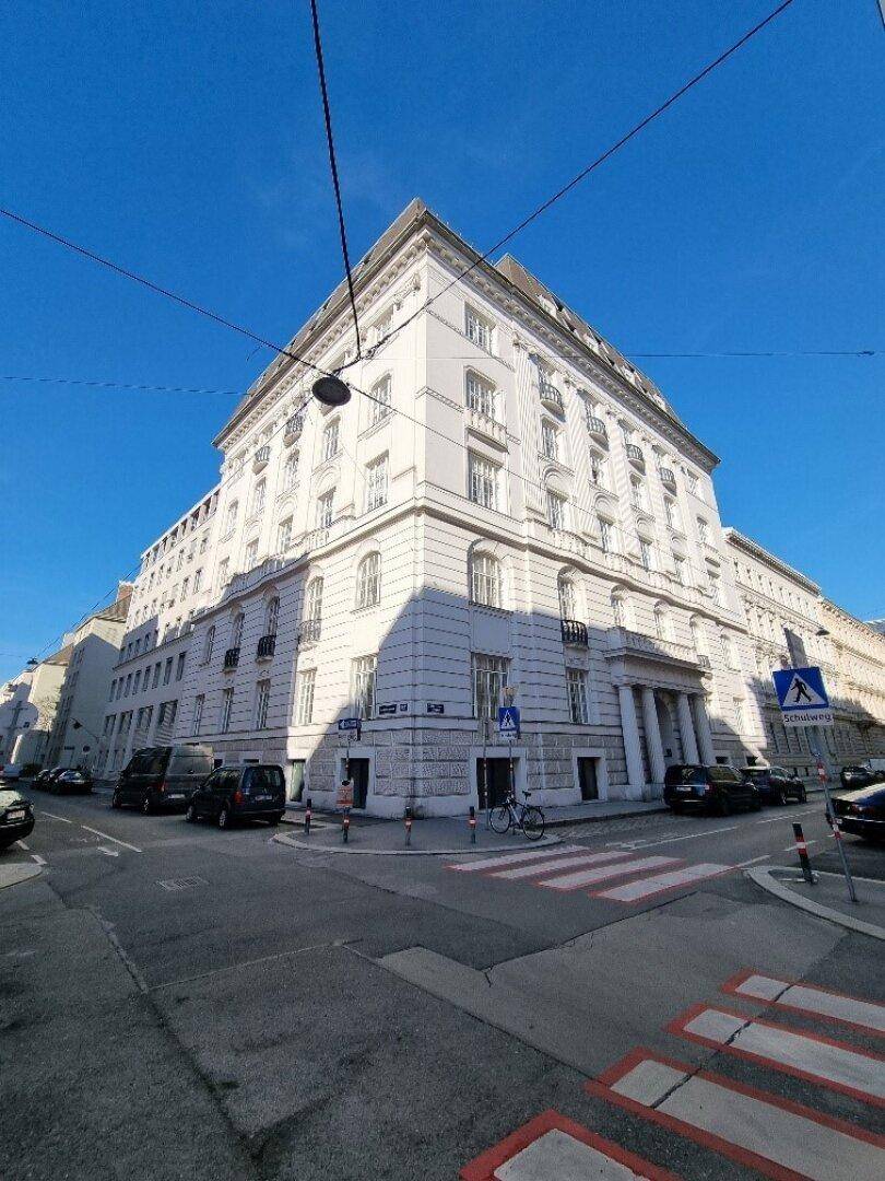 Großes, weißes Eckgebäude mit klassischer Architektur und vielen Fenstern unter blauem Himmel.