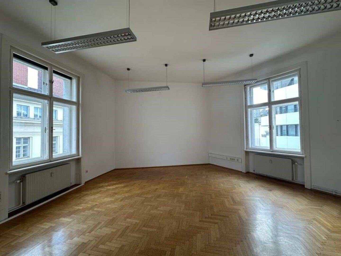 Heller und leerer Raum mit klassischem Fischgrätparkett und zwei großen Fenstern zur Straße.