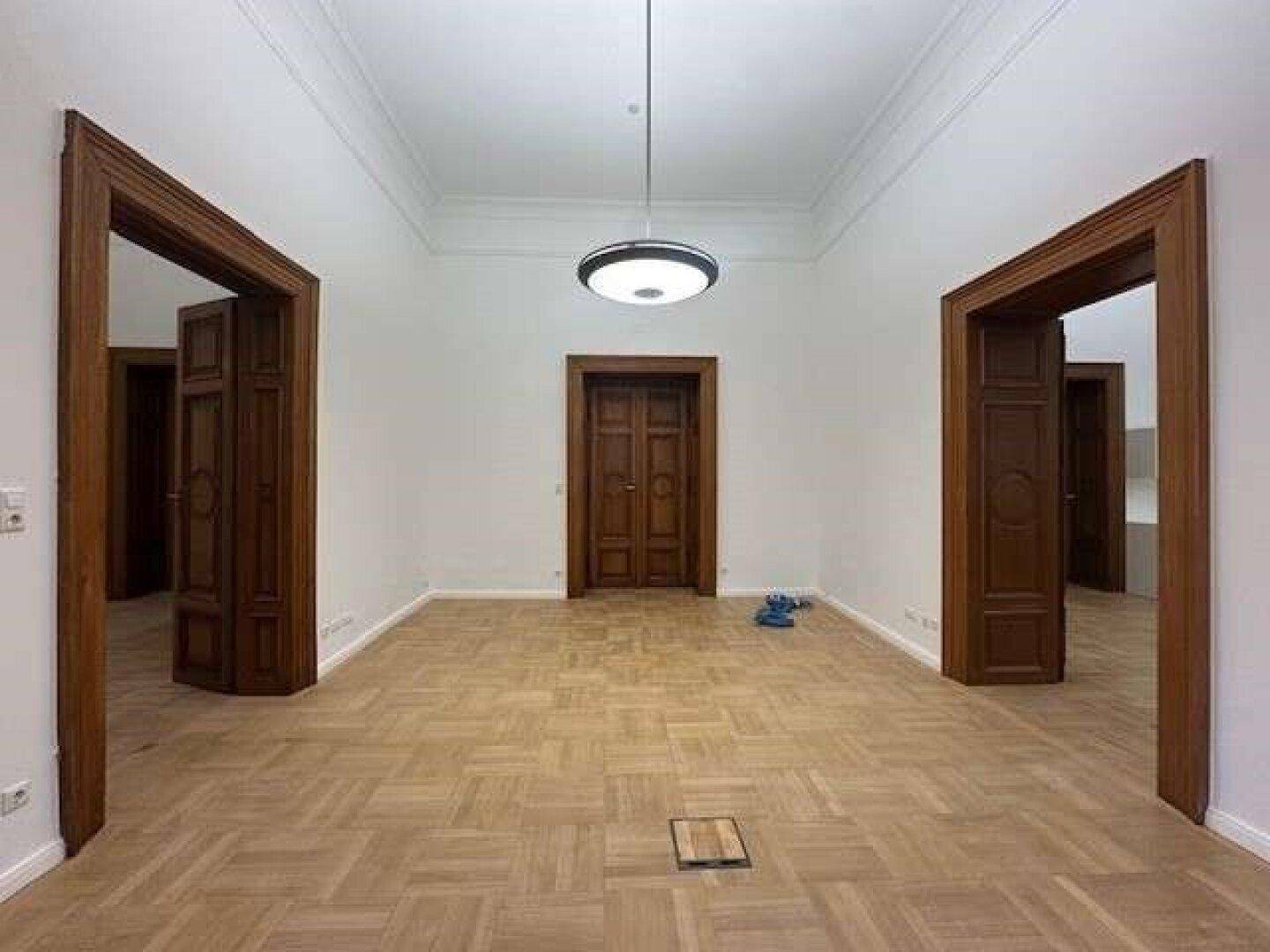Leerer Raum mit hellem Parkettboden, hohen Decken und klassischen Holztüren in einem Altbau.