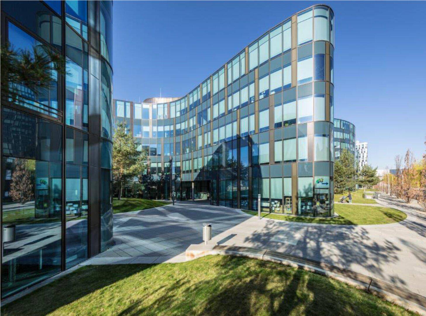 Eingangsbereich eines modernen Bürokomplexes mit geschwungener Glasfassade und gepflegten Grünflächen.