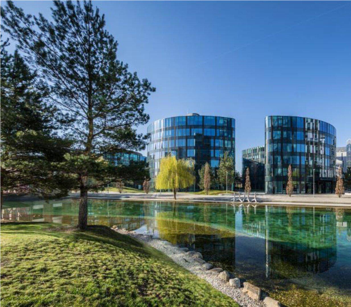 Mehrere moderne Bürogebäude mit Glasfassaden, umgeben von Wasserflächen und Bäumen unter blauem Himmel.