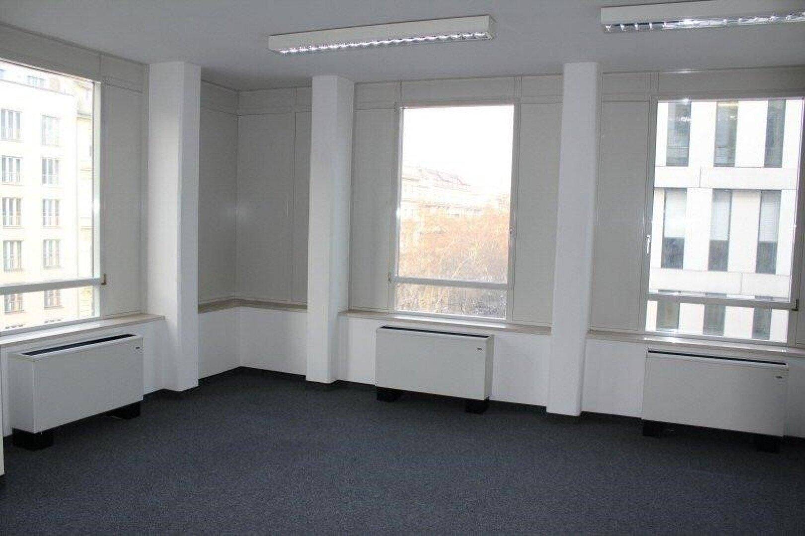 Heller Büroraum mit großen Fenstern, die einen Blick auf die Stadt und angrenzende Gebäude bieten.